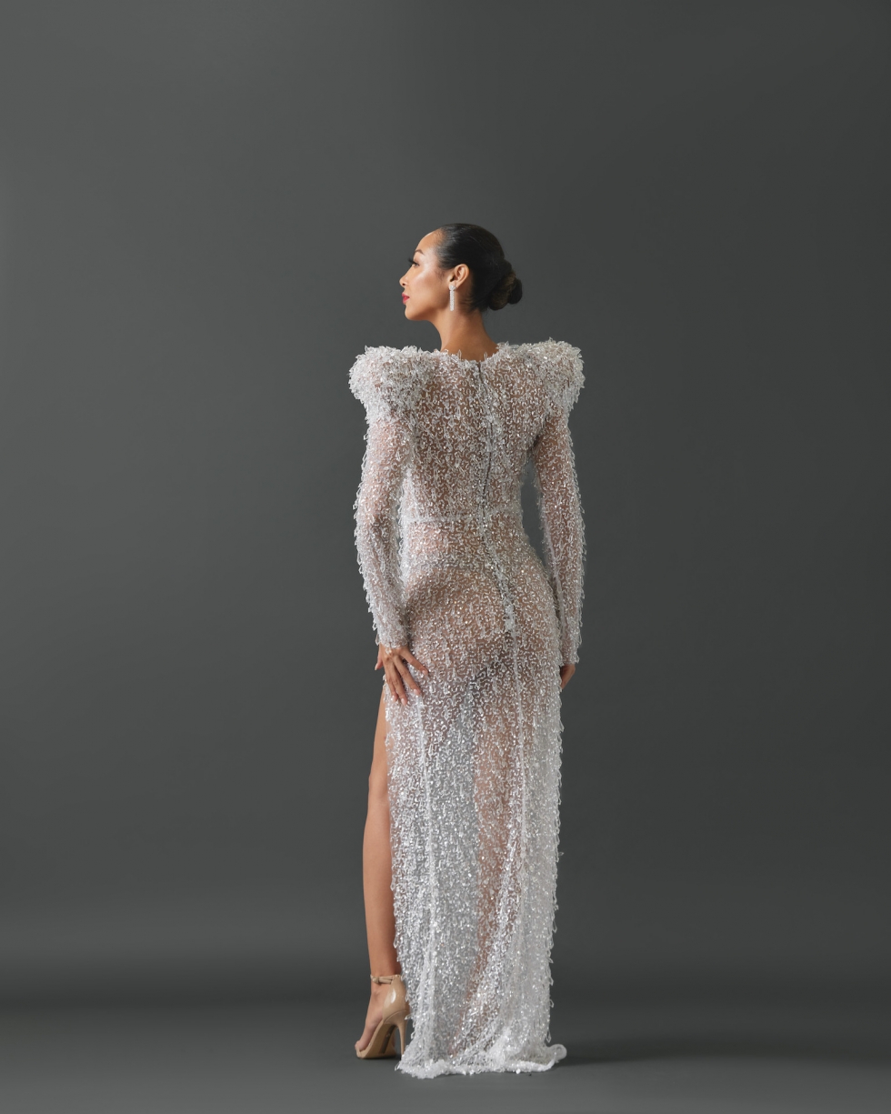 Sheer column gown with a high slit and deep V in neckline. Gown features long sleeves and exaggerated, structured shoulders. Fabrication and embellishment of gown features heavily patterned sequin mesh and covered with large prism shaped, hanging glass beads.