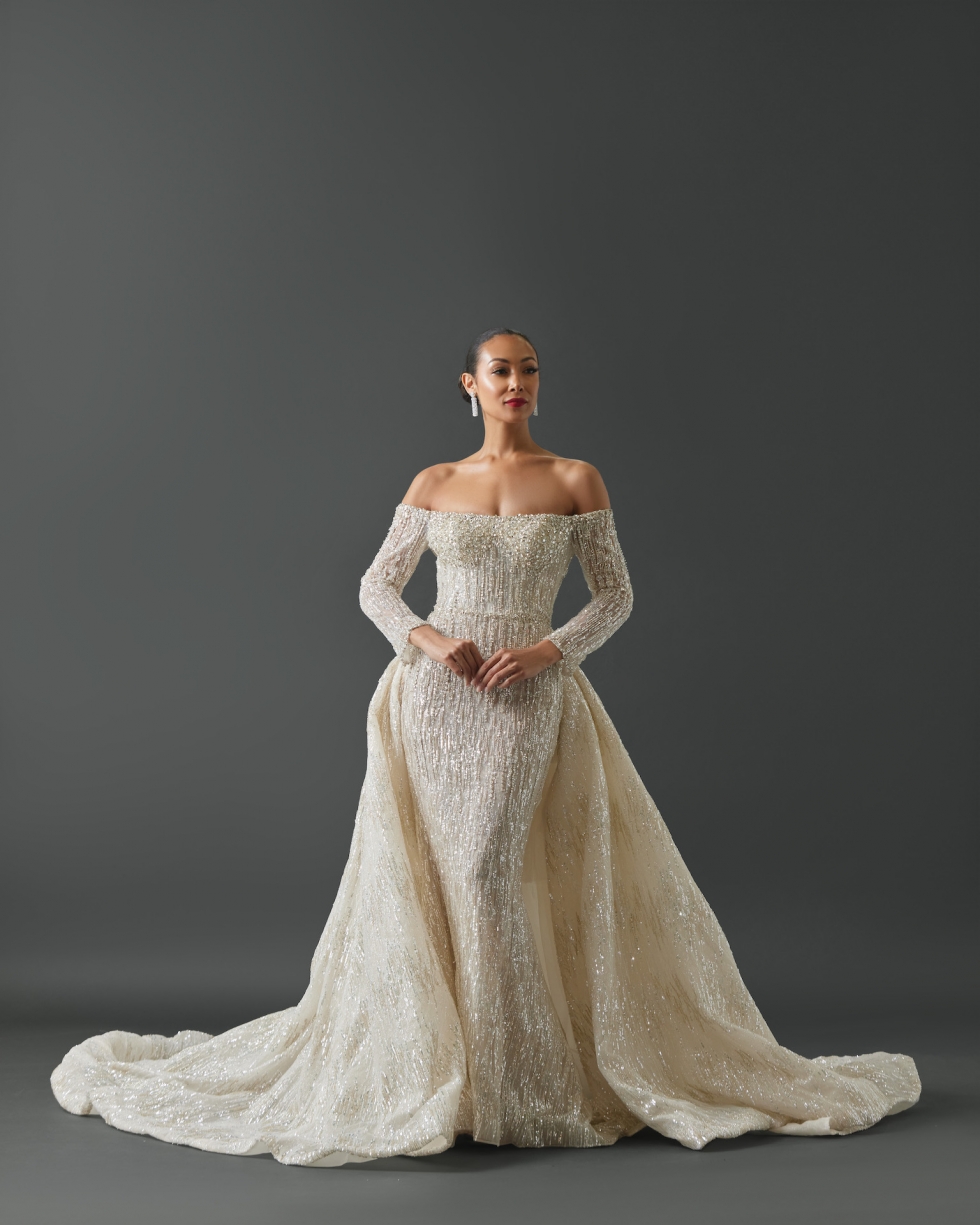 Long sleeve off shoulder fit and flare gown. Gown features lined sequin and crystal pattern heavily throughout gown with larger crystals framing neckline. Overskirt attached at waist from the side seam comprised of same fabrication and embellishments.